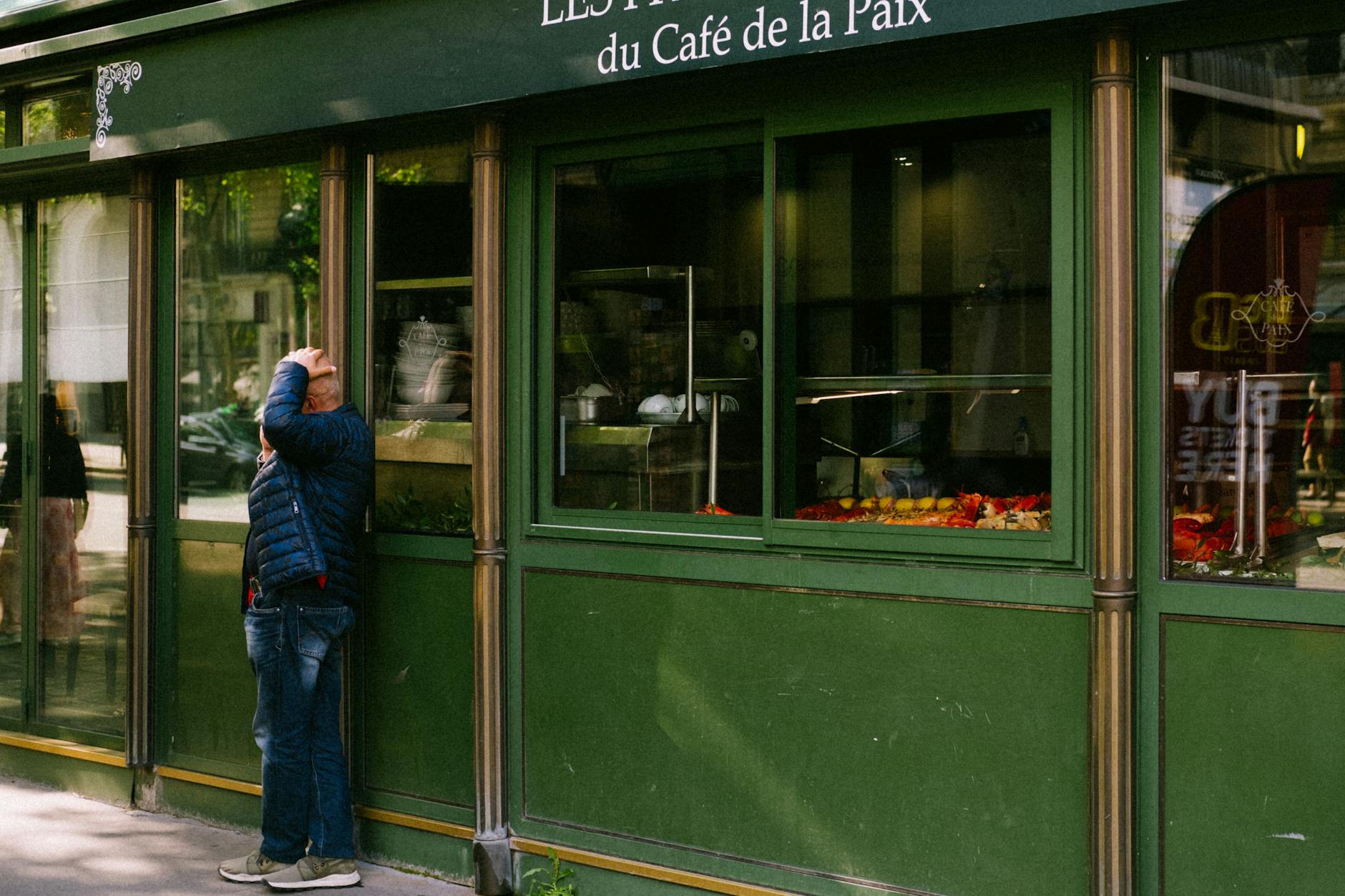 A man stands outside a green café with glass windows in an urban area, reflecting street life.
