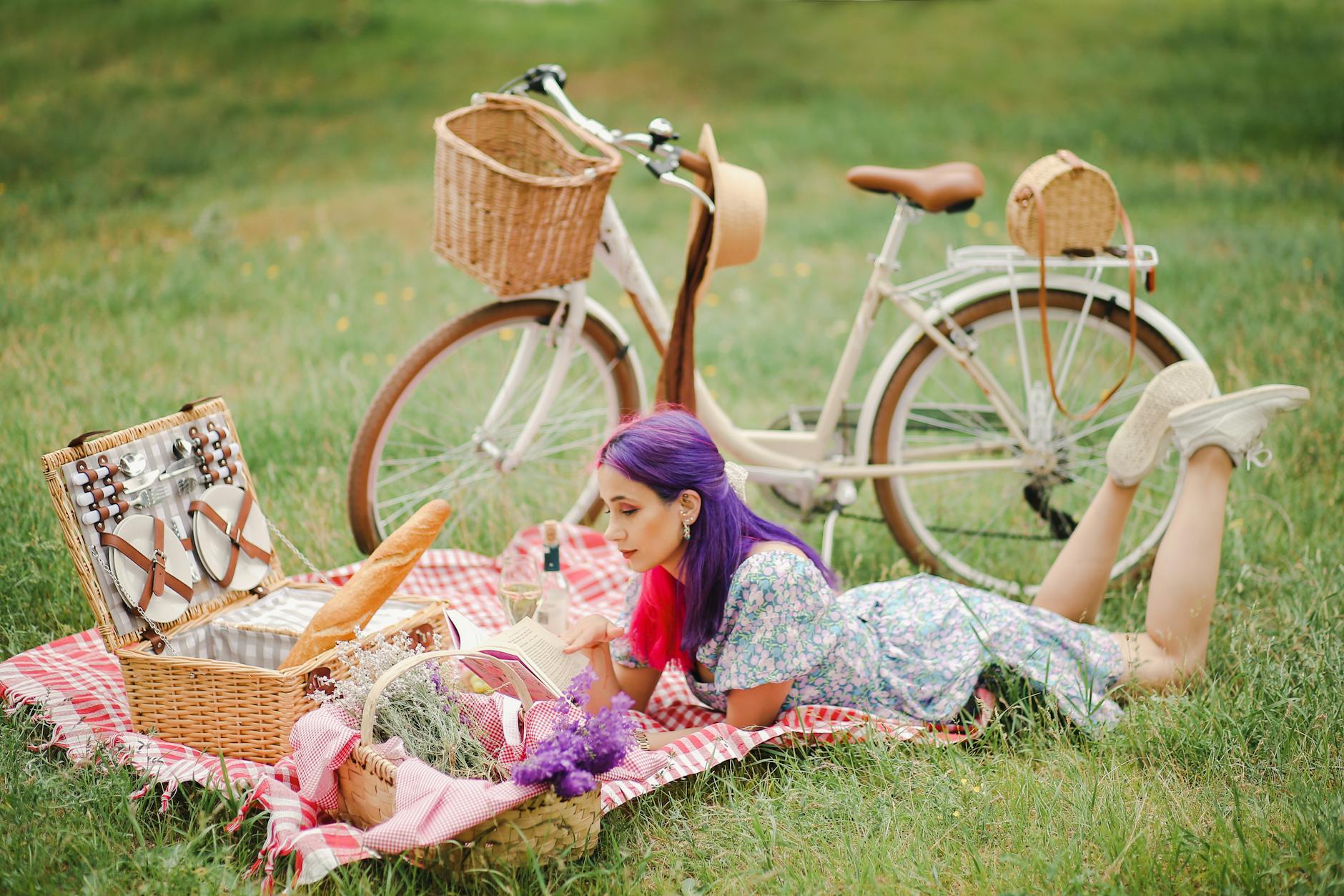 A woman lies on a picnic blanket with a bicycle and basket, reading in a sunny outdoor setting.
