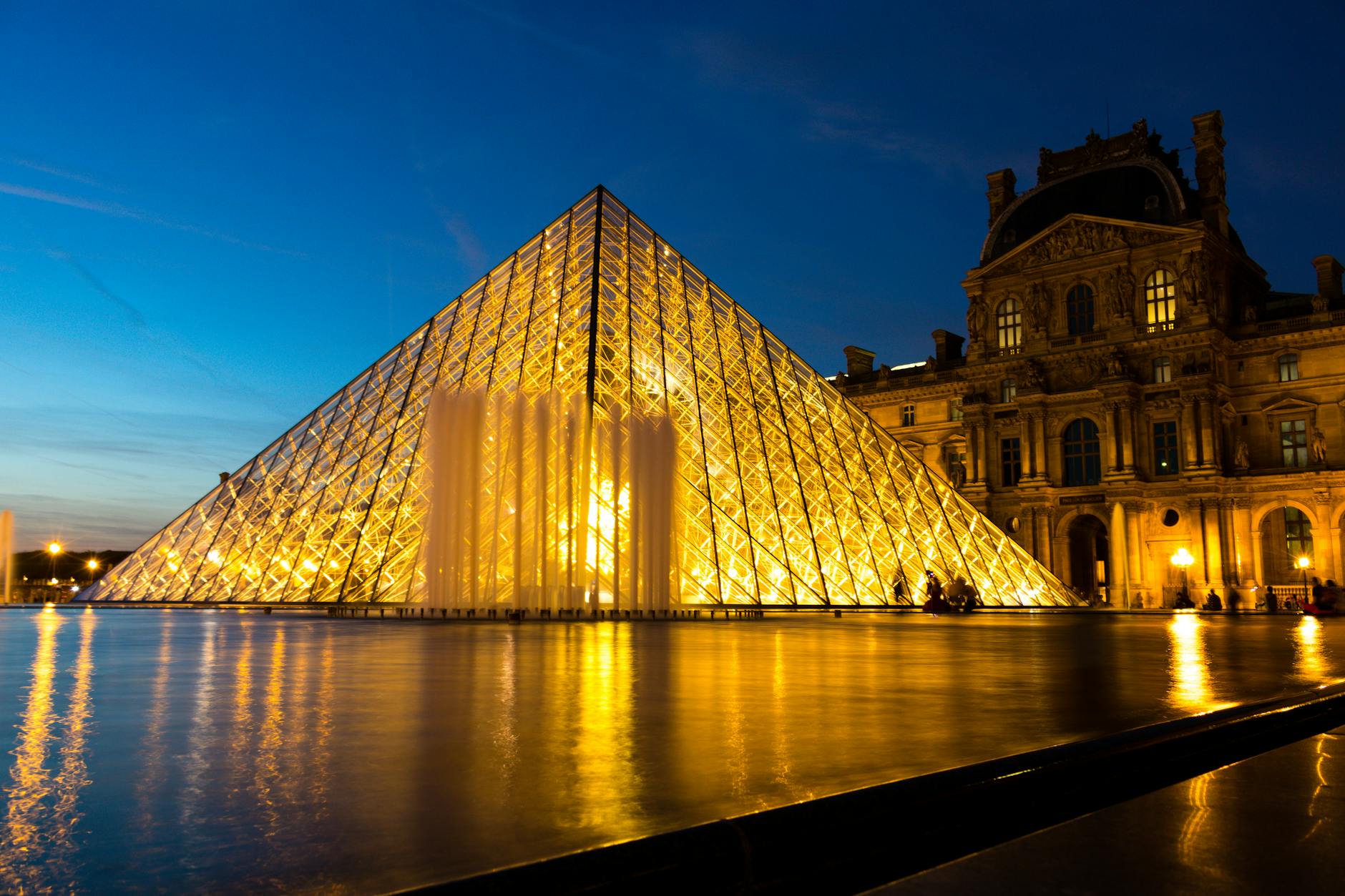The Louvre Pyramid illuminated beautifully in the evening, reflecting in the courtyard's water.