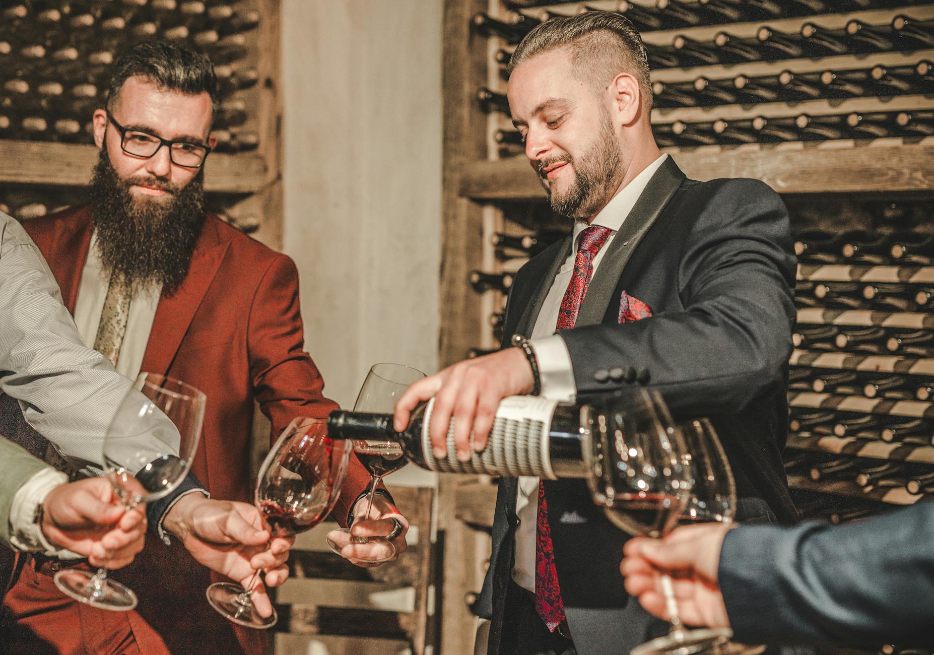 Two men in suits share wine at a stylish cellar gathering.