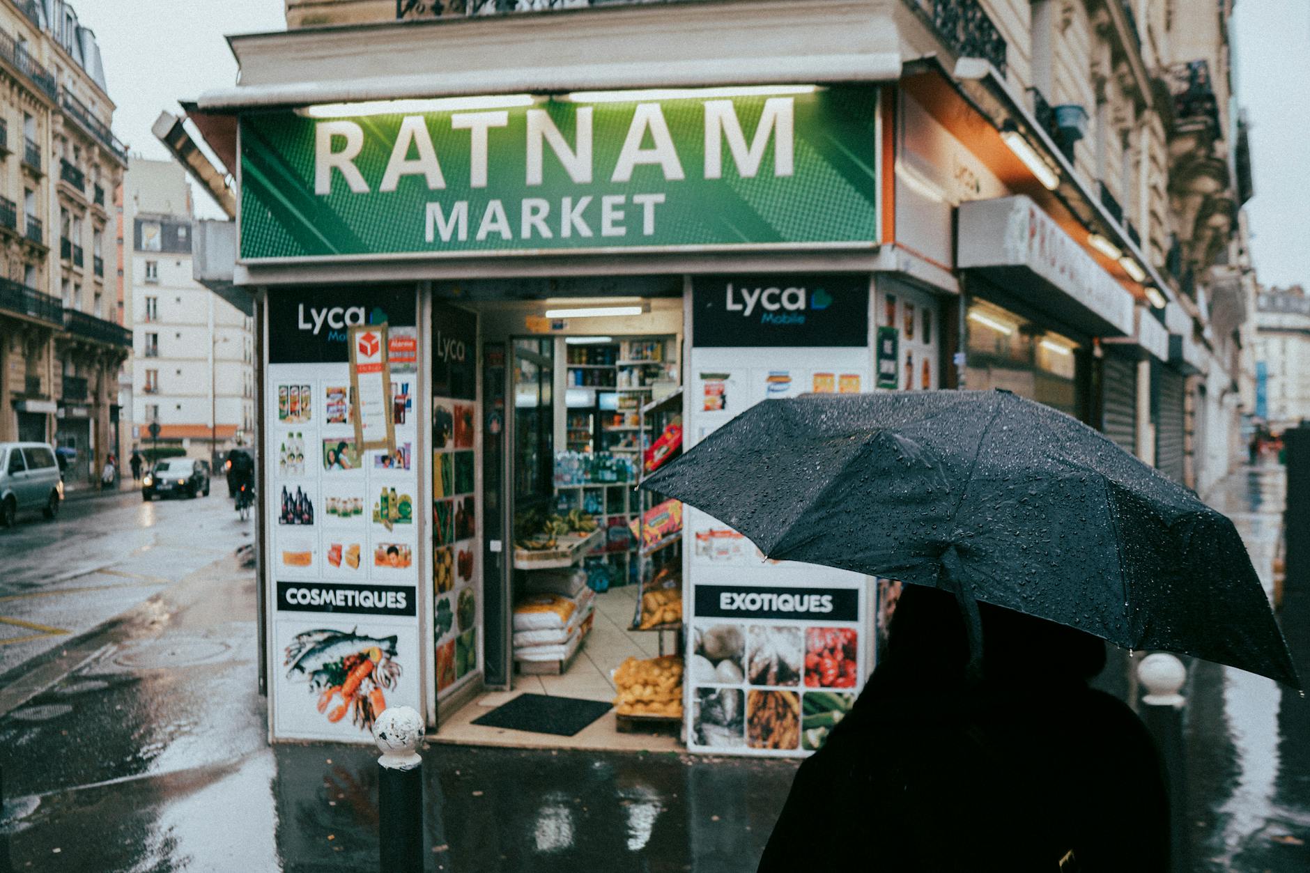 A rainy day in Paris featuring a small corner market with a pedestrian holding a black umbrella.