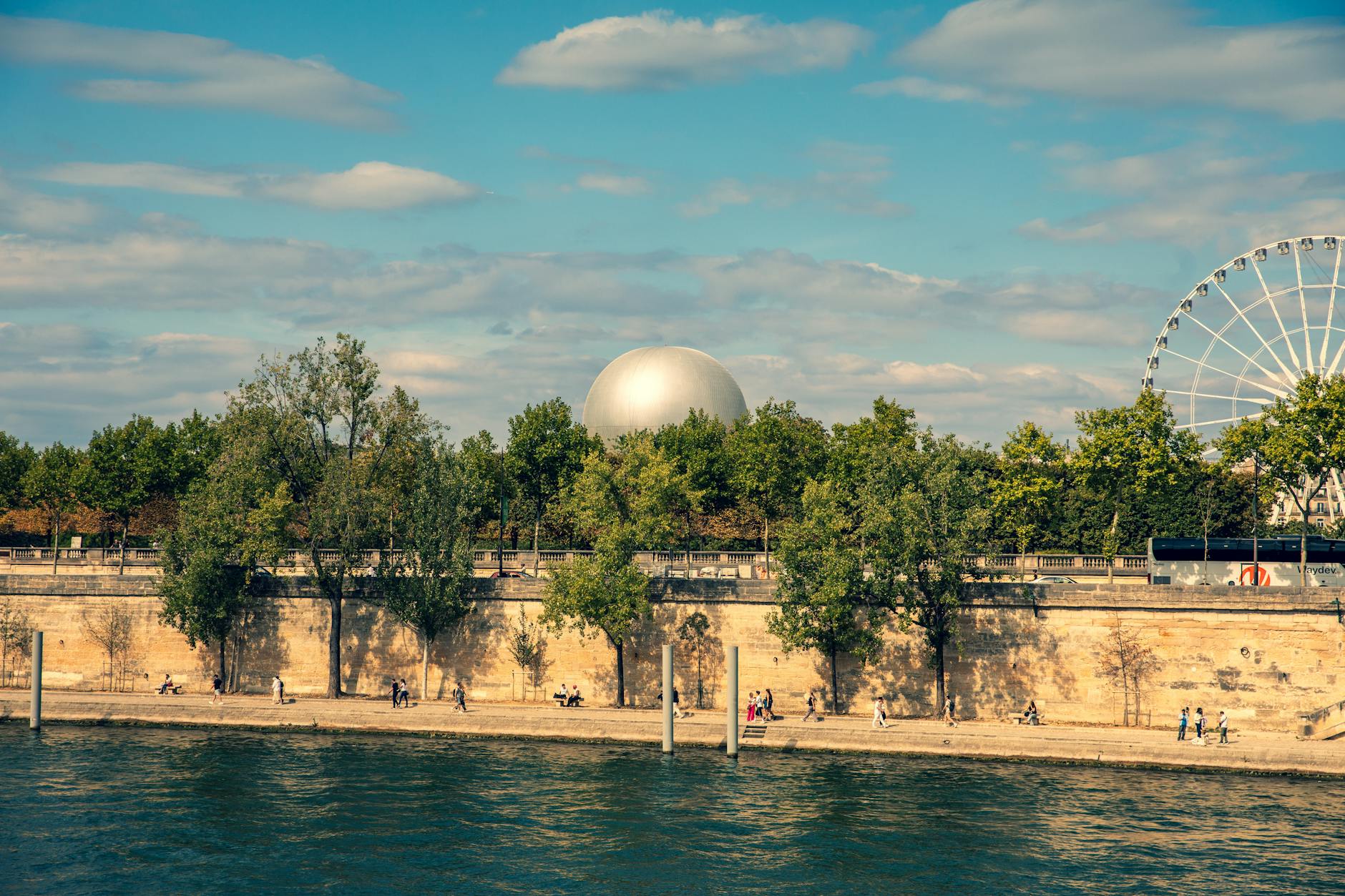 Picturesque view of a sunny Paris riverbank with trees, a Ferris wheel, and dome.