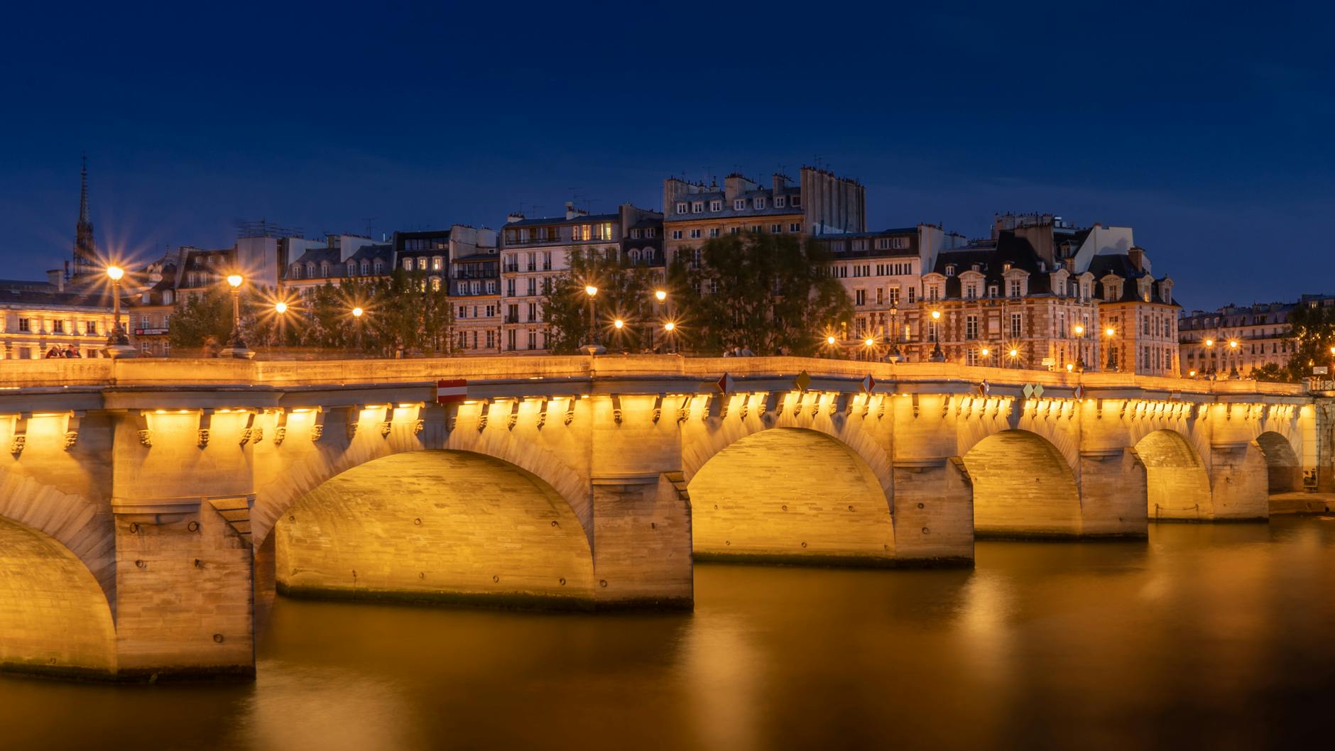 Illuminated Pont Neuf bridge reflecting on the Seine River at night in Paris, creating a serene cityscape.