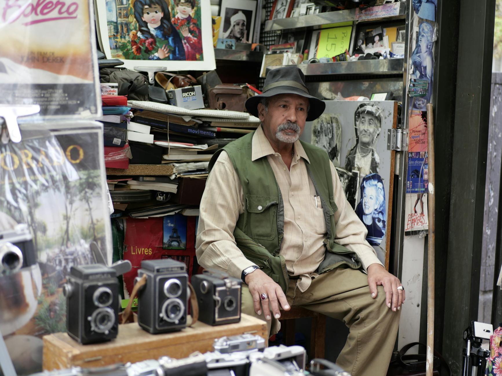 Elderly man at a Parisian market stall selling vintage cameras and artwork.