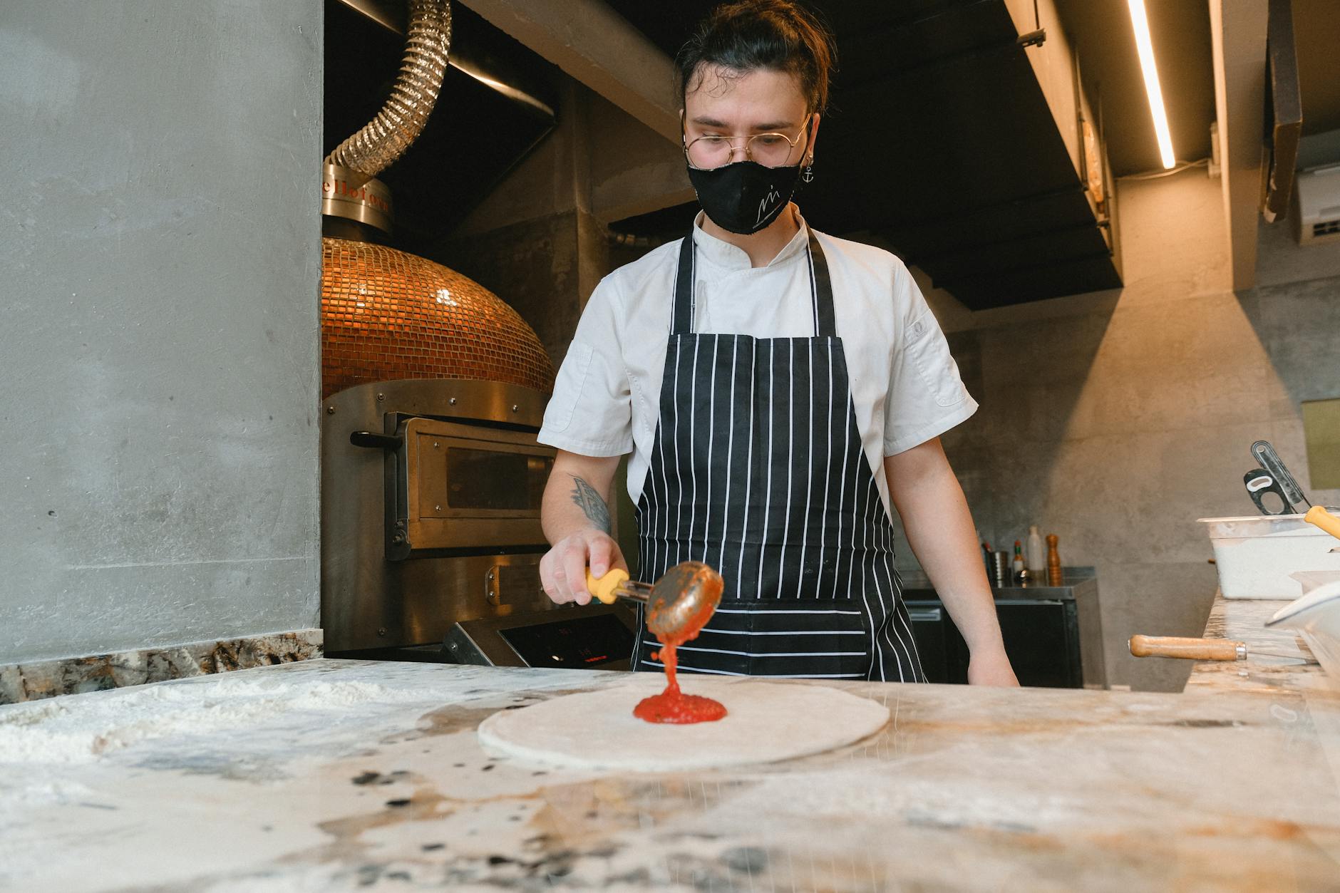 Chef wearing mask spreads tomato sauce on pizza base in kitchen.