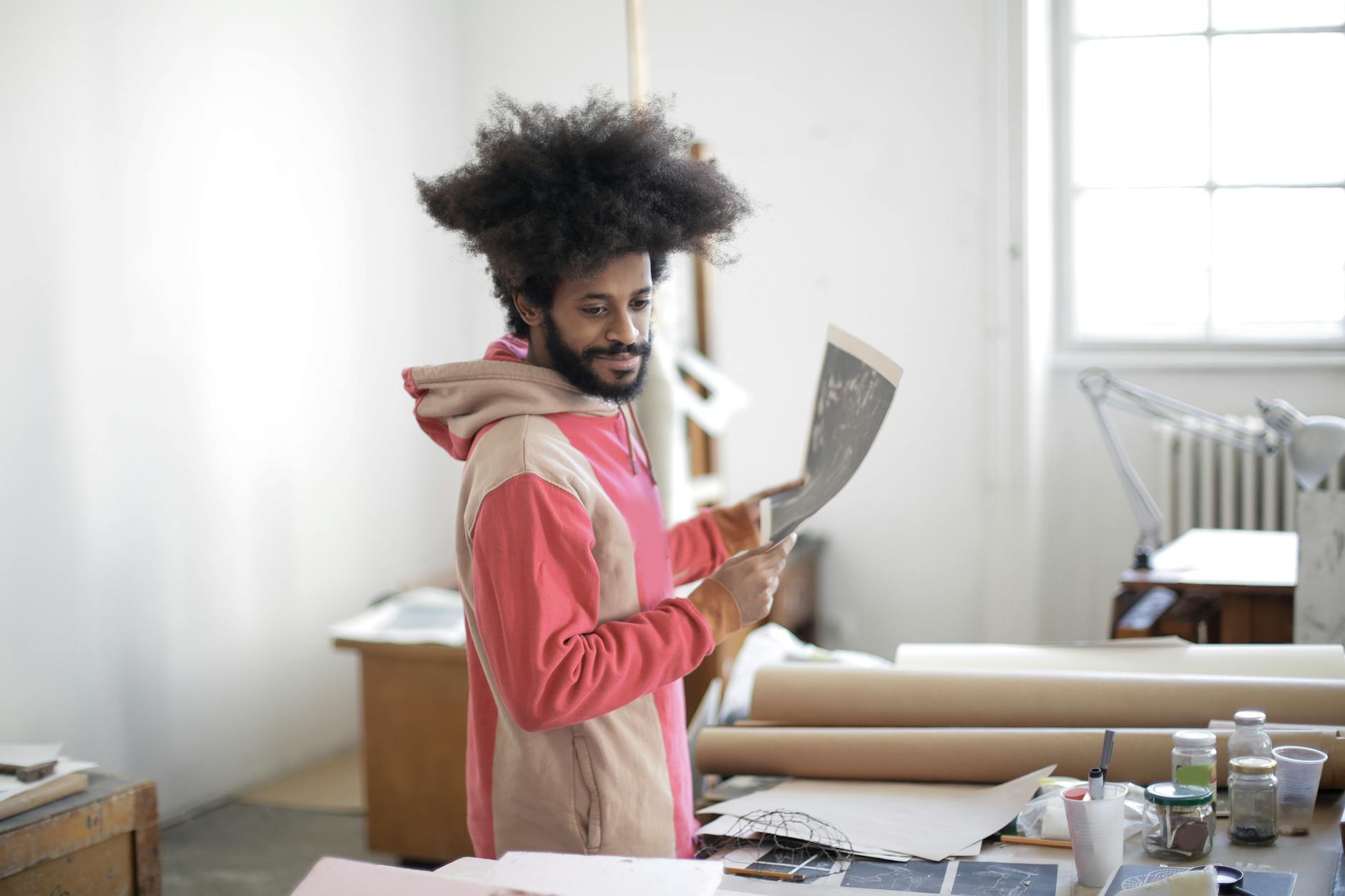 Young bearded ethnic male master with creative Afro hairstyle wearing hoodie sweatshirt standing with printed samples in hands near table with craft paper and dyes in aged atelier with shabby furniture