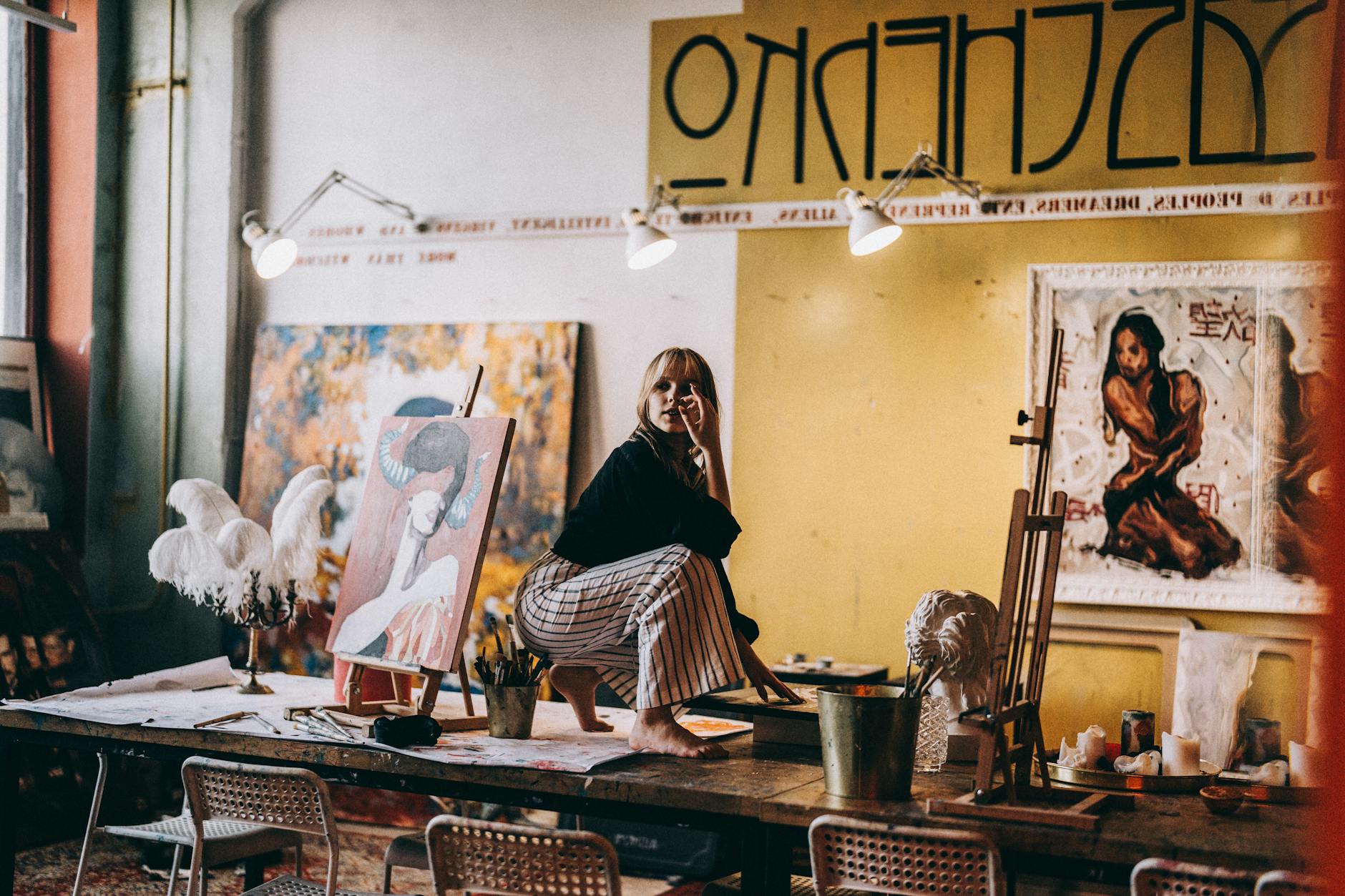 A woman artist sitting on a work table in a vibrant art studio surrounded by paintings and sculptures.