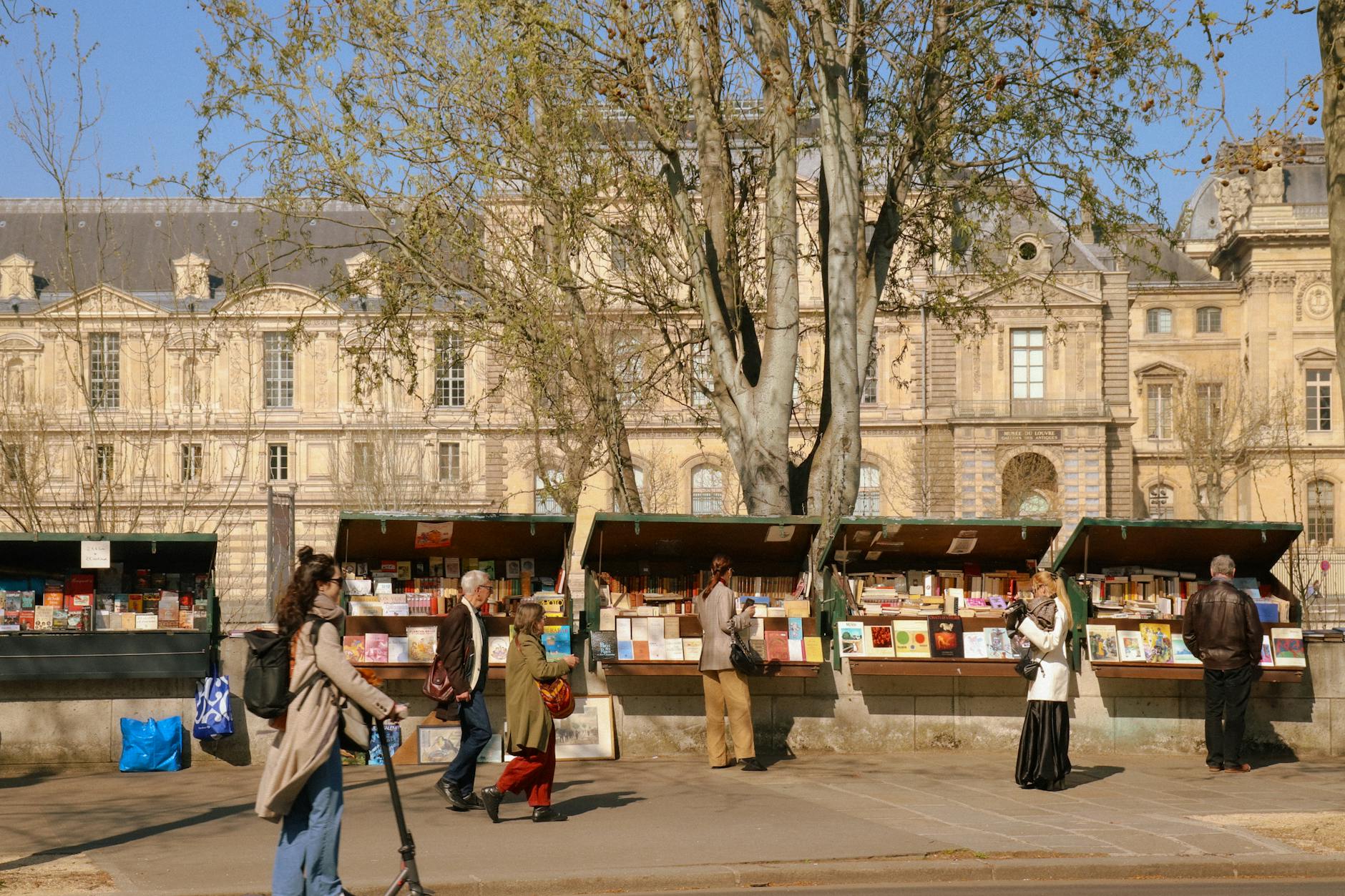 People browsing bookstalls by the Seine River in Paris on a sunny day.