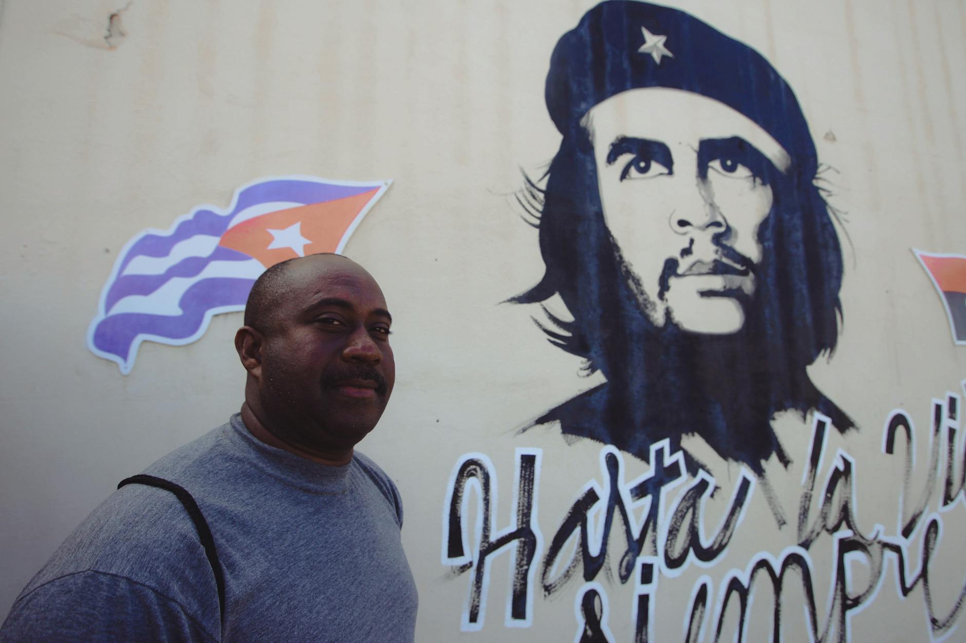 African American man with Che Guevara mural and Cuban flag in the background, outdoor setting.
