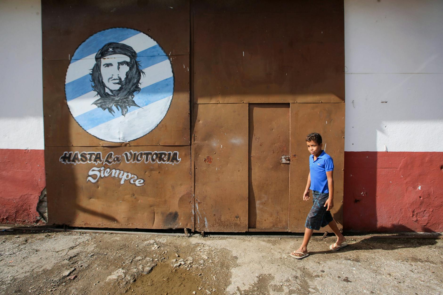A child walks past a street mural of Che Guevara, featuring vibrant artwork and a metal gate.