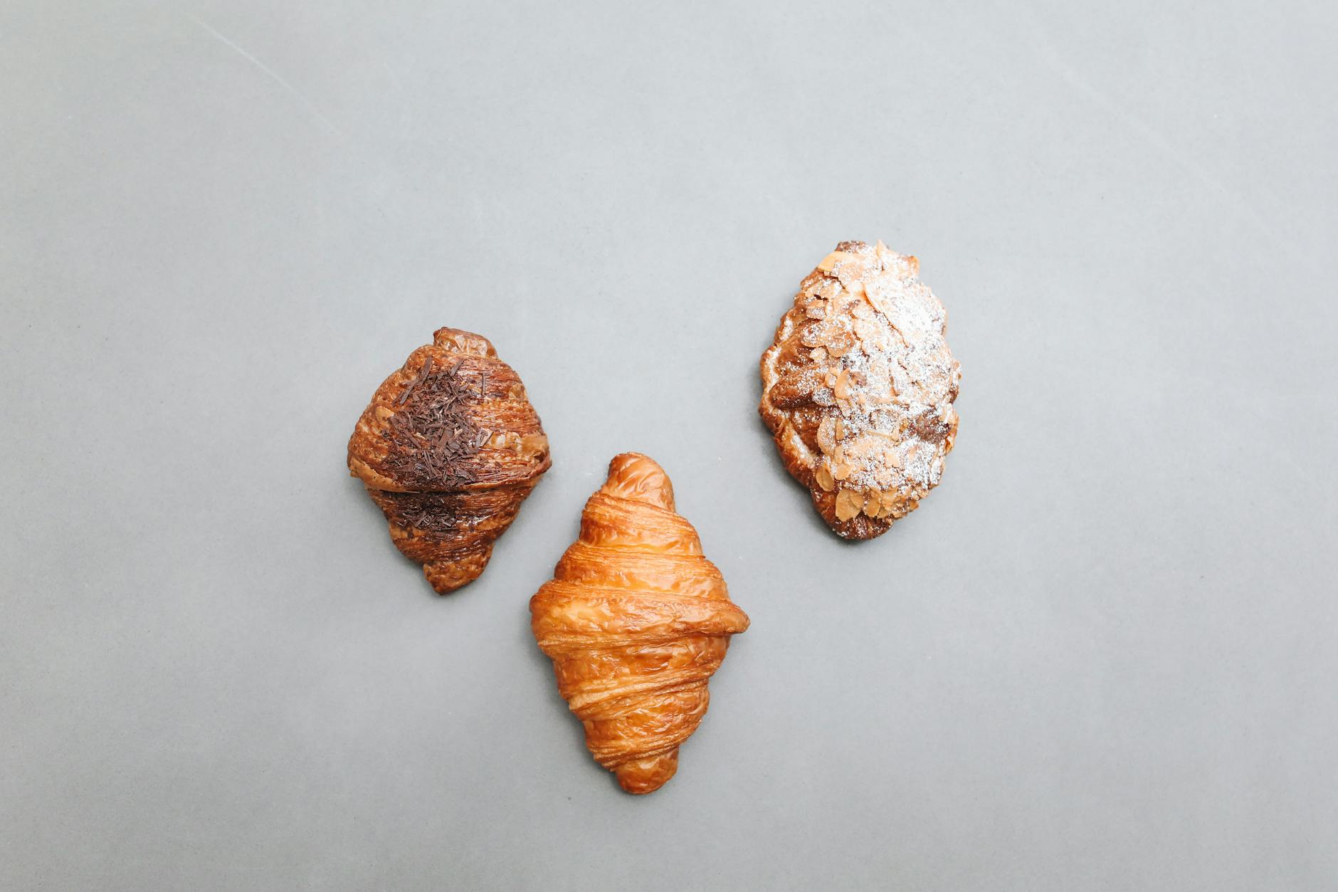 A trio of artisan croissants including plain, chocolate, and almond on a gray background in a flat lay style.