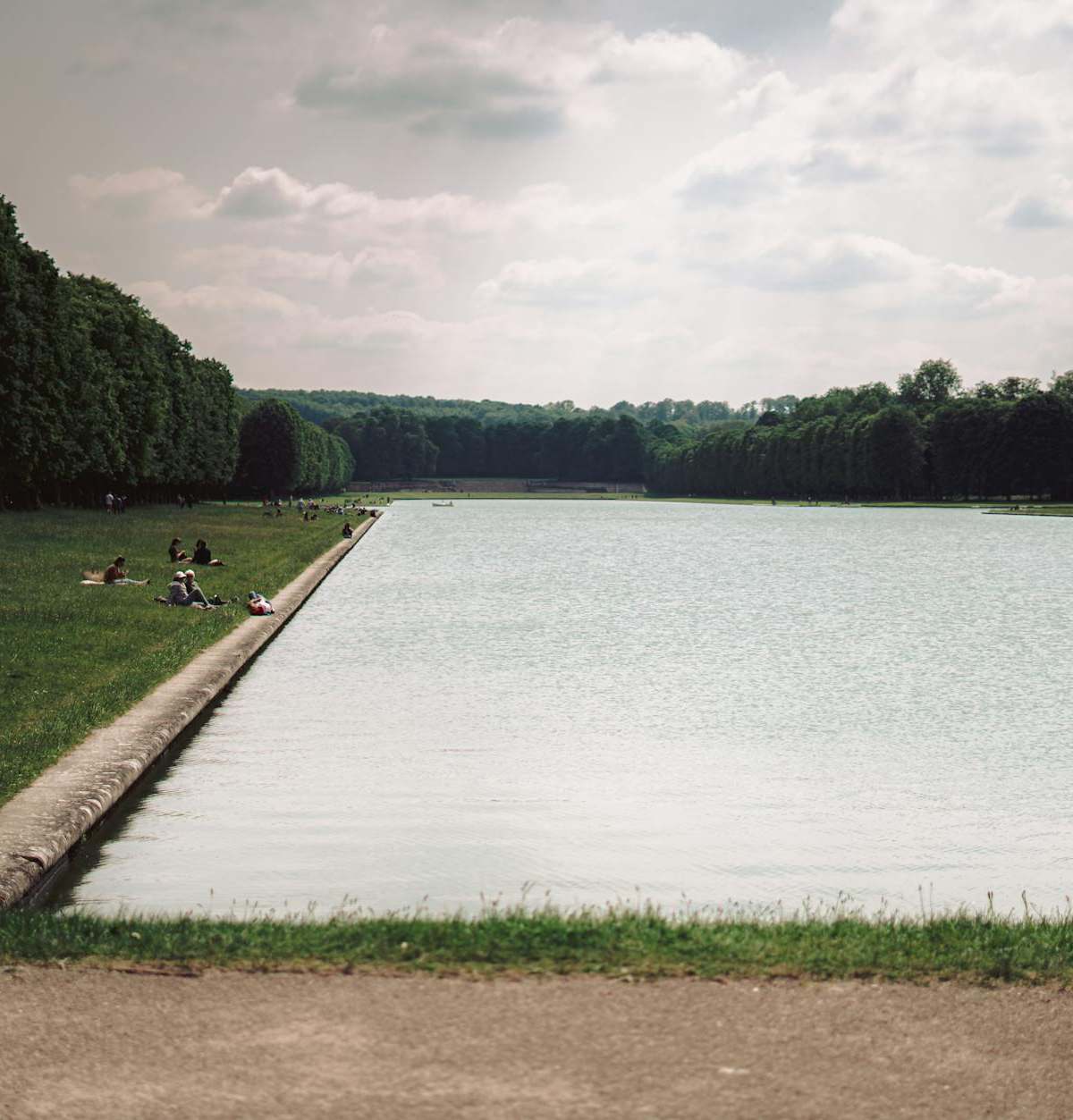 People enjoying a sunny day by the canal in the gardens of Versailles.