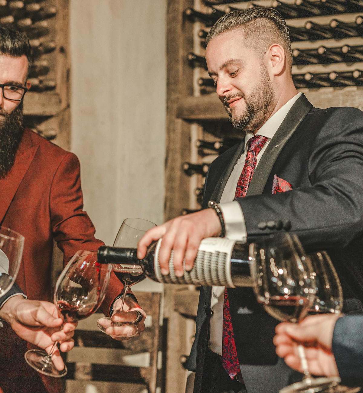 Two men in suits share wine at a stylish cellar gathering.