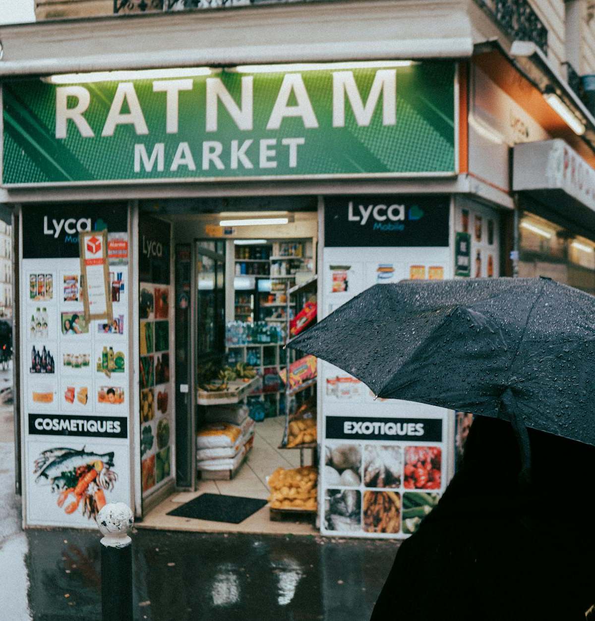 A rainy day in Paris featuring a small corner market with a pedestrian holding a black umbrella.