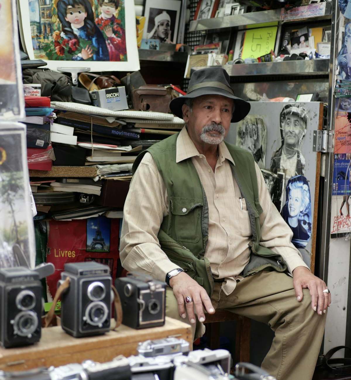 Elderly man at a Parisian market stall selling vintage cameras and artwork.